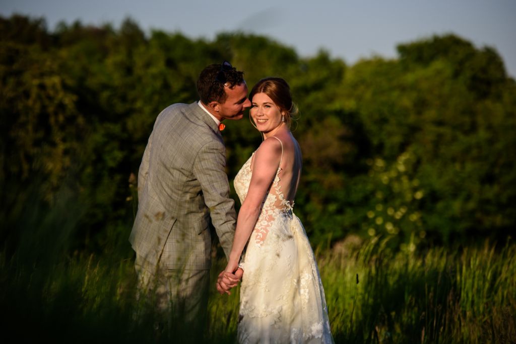 Groom kissing bride in long grass at sunset at Monkton Barn